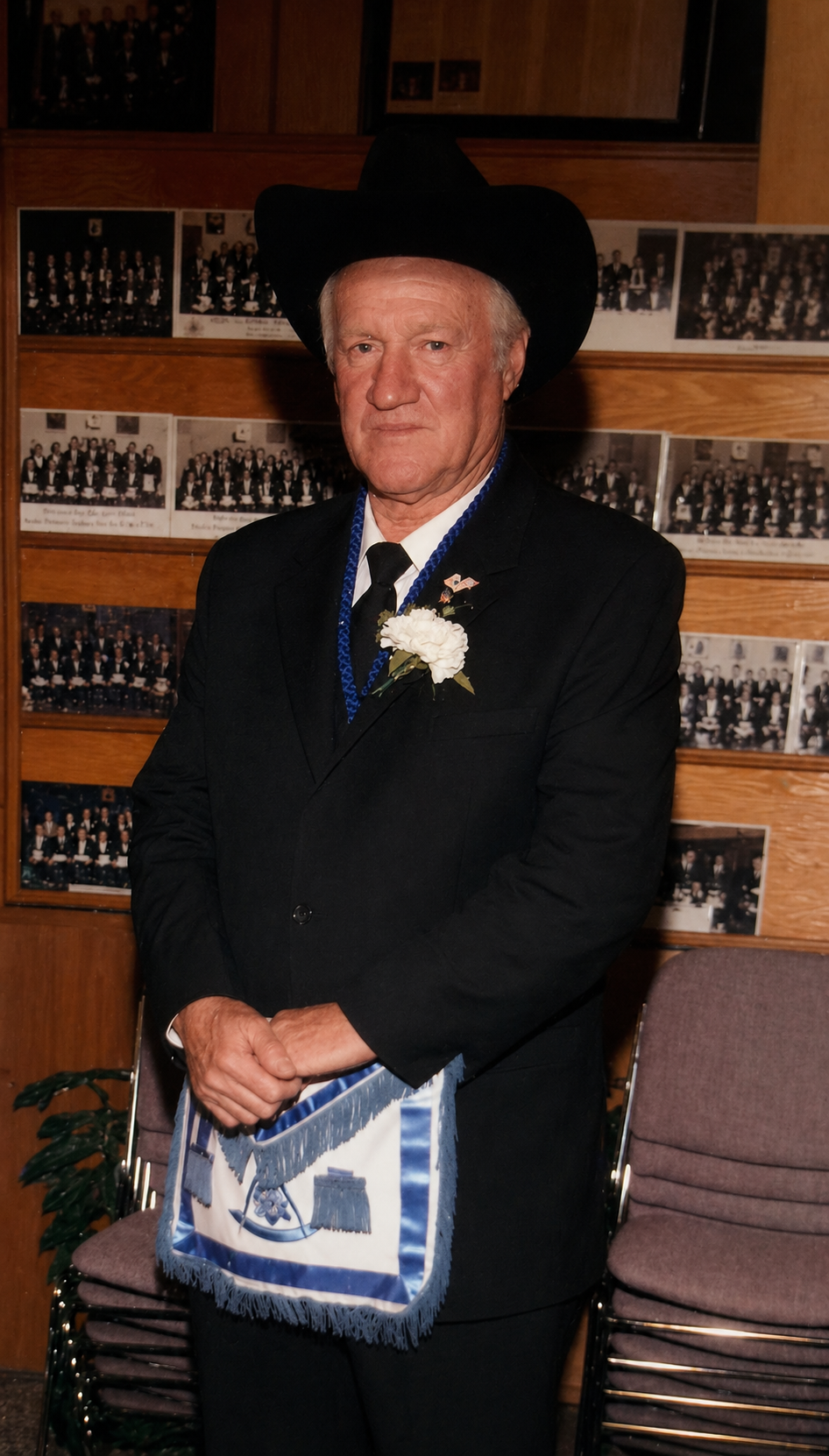 Brother in black suit and cowboy hat wearing a blue and white Masonic apron, standing before a wall of lodge portraits