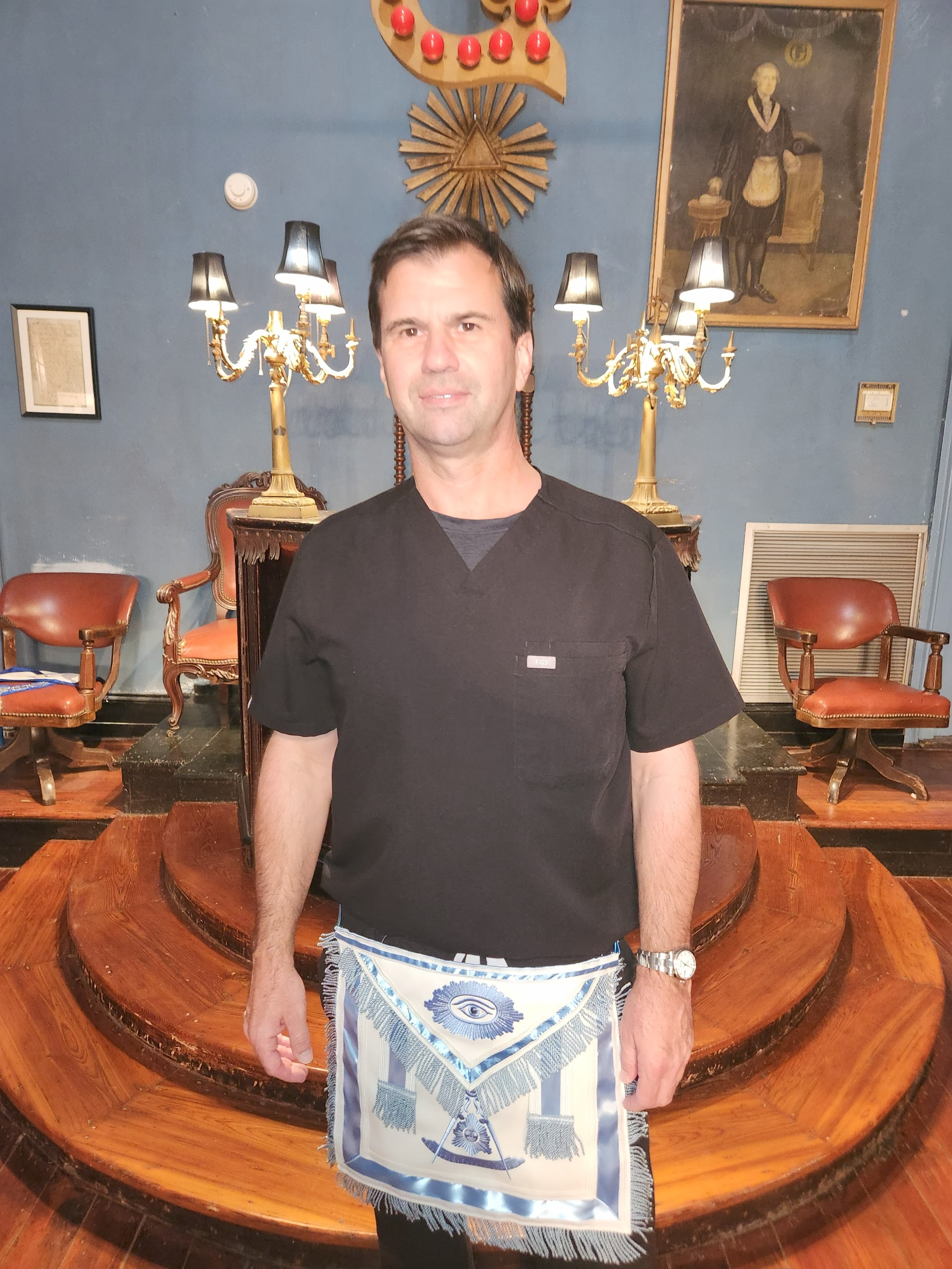 Brother in black scrubs wearing an ornate blue and white Masonic apron standing in the lodge hall