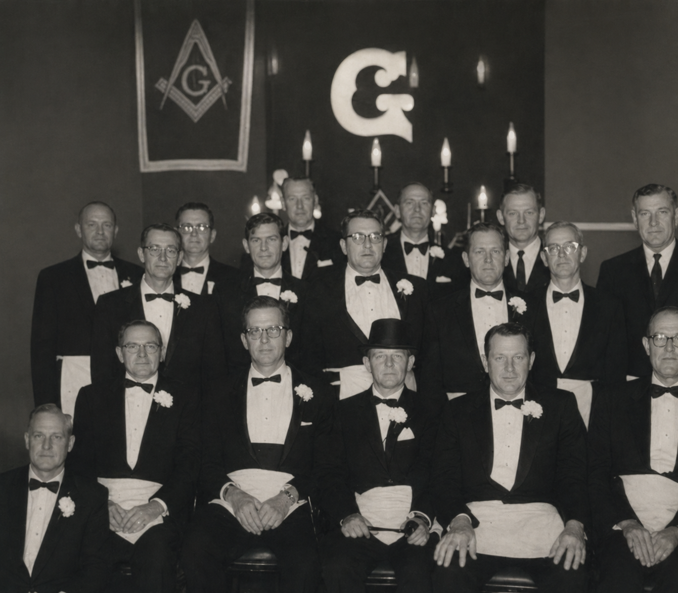 Lodge officers in tuxedos and Masonic aprons posed beneath the square and compass symbol with candelabra, 1950s